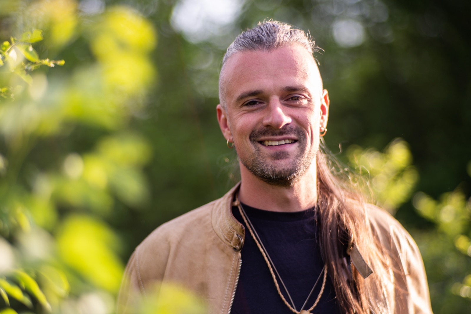 Portrait of Axel Drioli smiling, in a tan jacket against green woodland
